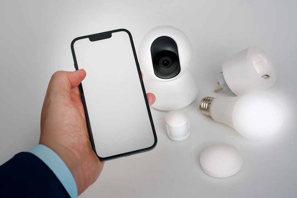Eine Hand hält ein Smartphone, dahinter eine schwenkbare Kamera für innen und weitere Smart-Home-Geräte.