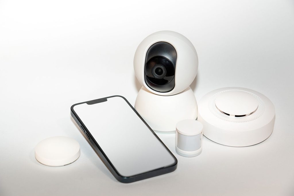Weiße WLAN-Kamera für innen neben einem Smartphone und kleinen Funksensoren auf hellem Untergrund.