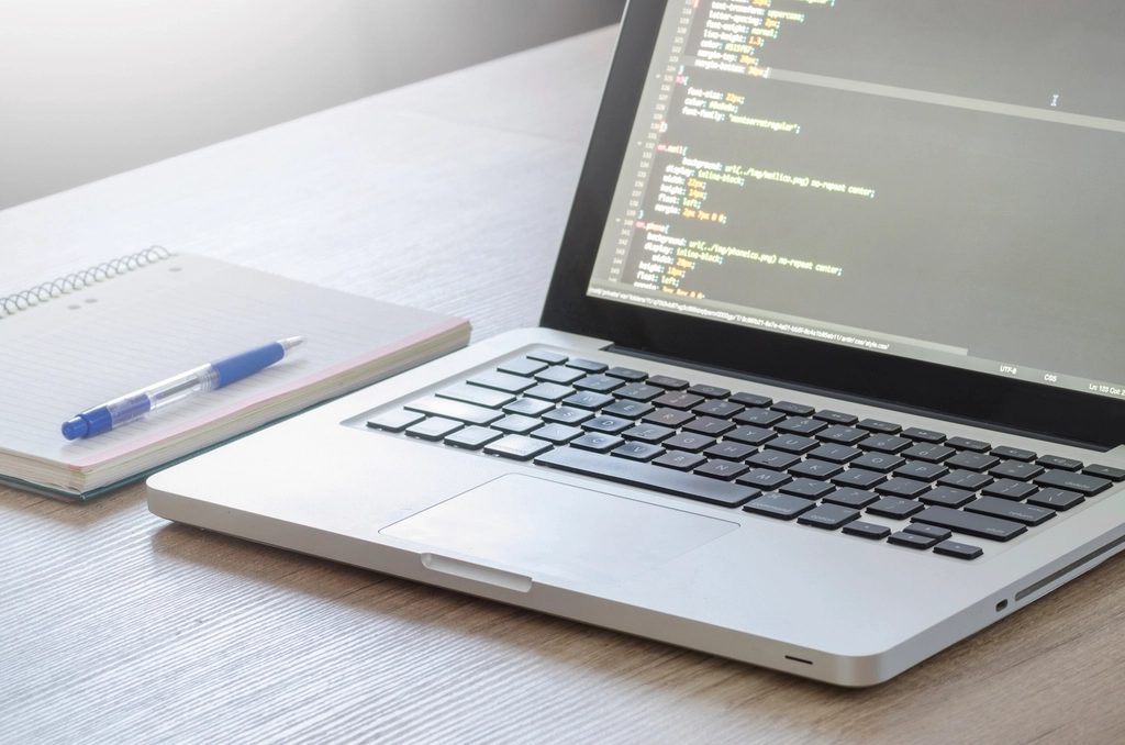 Aufgeklappter Laptop mit CSS-Code im Editor auf einem hellen Holztisch, daneben ein Notizblock mit Kugelschreiber.