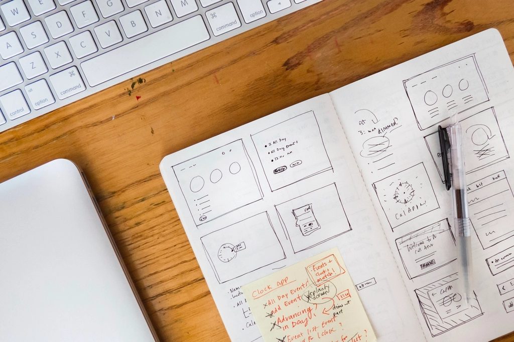 Aufgeschlagenes Skizzenbuch mit handgezeichneten Wireframes für Website-Layouts, daneben Tastatur, Laptop und Stift auf einem Holztisch.